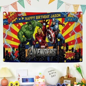 May include: A colorful Marvel Avengers birthday banner with Hulk, Iron Man, and Captain America. The banner has the text "Happy Birthday Jason" and "Avengers" with comic book sound effects. Party decorations are in the foreground.