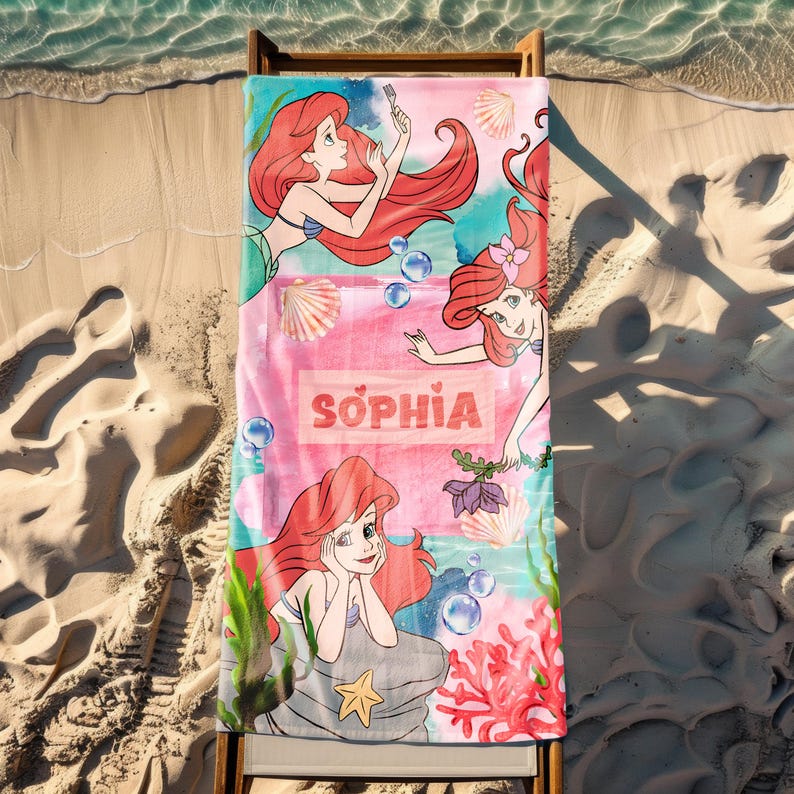 May include: A beach towel featuring the character Ariel from The Little Mermaid. The towel has a pink and teal background with Ariel illustrations, seashells, bubbles, and the name "Sophia" in a rectangular box. The towel is lying on a wooden beach chair on the sand.