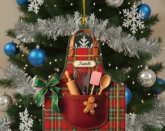 May include: A Christmas tree ornament shaped like a red plaid apron with a pocket. The apron features a gingerbread man, rolling pin, whisk, spatula, and spoon. The name "Isabelle" is printed on the apron. A green bow is attached to the apron.
