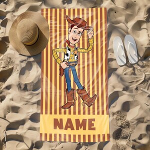 May include: A yellow and brown striped beach towel with a cartoon image of Woody from Toy Story. Woody is wearing his signature cowboy hat and vest. The towel has the word "NAME" printed on it in brown letters.