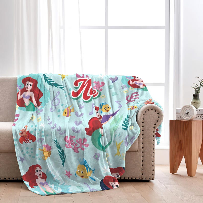 Puede incluir: Una manta azul claro con un dise&ntilde;o de sirena. La manta presenta ilustraciones de sirenas, peces y notas musicales. La palabra "Me" est&aacute; escrita en rojo y blanco. La manta est&aacute; sobre un sof&aacute; beige.