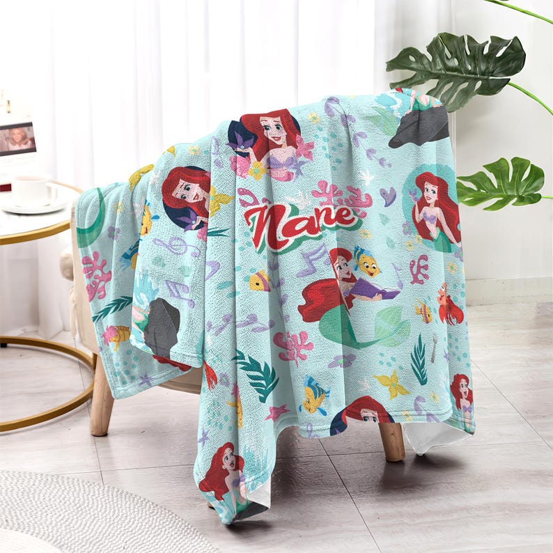 Puede incluir: Una manta azul claro con el personaje Ariel de La Sirenita. La manta tiene un patr&oacute;n repetido de Ariel, notas musicales y criaturas marinas. El nombre "Kane" est&aacute; escrito en escritura roja en la manta.