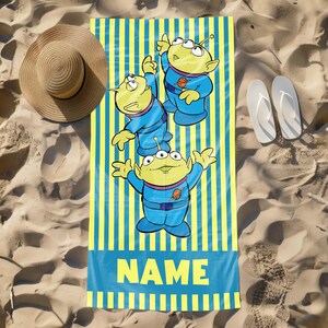 May include: A yellow and blue beach towel with a striped pattern. Three green aliens with three eyes each are featured on the towel. The towel has the word "NAME" printed on it.