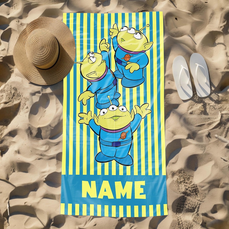 May include: A yellow and blue beach towel with a striped pattern. Three green aliens with three eyes each are featured on the towel. The towel has the word "NAME" printed on it.