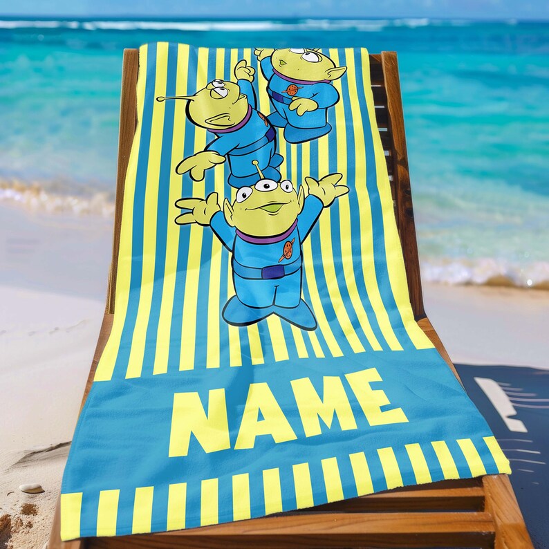 May include: A blue and yellow striped beach towel with three green aliens in blue space suits. The towel has the word "NAME" printed on it.