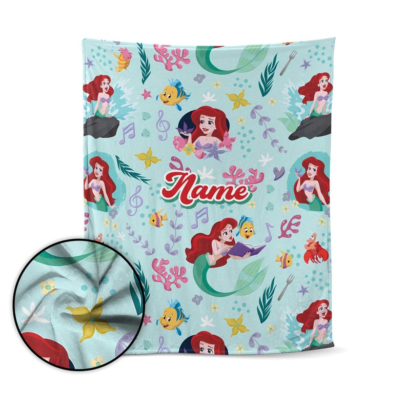 Puede incluir: Una manta azul claro con un patr&oacute;n repetido de una sirena con cabello rojo, notas musicales, peces y otros elementos tem&aacute;ticos del mar. La palabra "Name" est&aacute; escrita en cursiva en el centro de la manta.