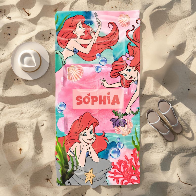May include: A beach towel featuring a colorful illustration of a mermaid with red hair. The towel has a pink background with the name "SOPHIA" in white letters. The design includes seashells, bubbles, and coral.