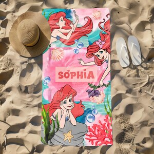 May include: A beach towel featuring the Little Mermaid, with the name "Sophia" in pink. The towel has illustrations of the mermaid, seashells, and aquatic plants. A straw hat and white flip-flops are on the sand next to the towel.