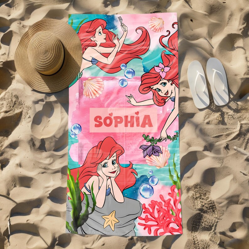 May include: A beach towel featuring the Little Mermaid, with the name "Sophia" in pink. The towel has illustrations of the mermaid, seashells, and aquatic plants. A straw hat and white flip-flops are on the sand next to the towel.