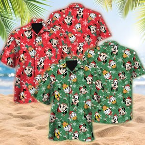 May include: Three short-sleeved button-down shirts with a festive pattern. The shirts are red and green, featuring cartoon characters like Mickey Mouse and Donald Duck in Santa hats and snowflakes. The shirts are displayed on a sandy beach with palm leaves in the background.