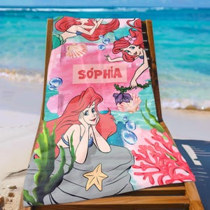 May include: A beach towel featuring a mermaid design with the name "SOPHIA" in pink. The towel has a pink and blue watercolor background with illustrations of seashells, coral, and seaweed. The mermaid has red hair and a blue top.