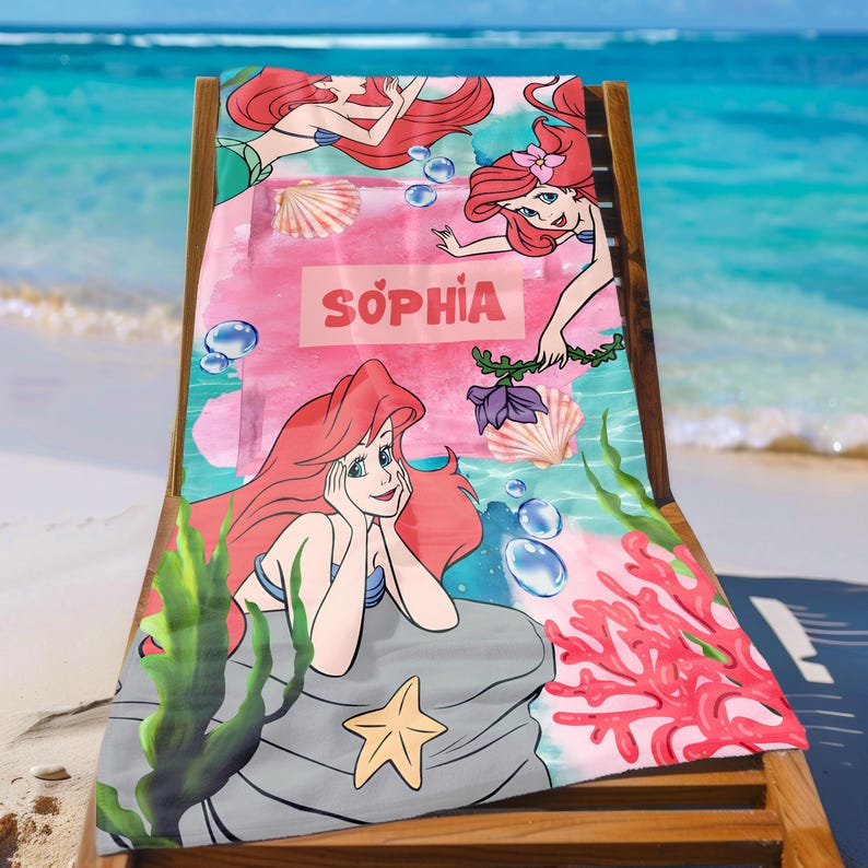 May include: A beach towel featuring a mermaid design with the name "SOPHIA" in pink. The towel has a pink and blue watercolor background with illustrations of seashells, coral, and seaweed. The mermaid has red hair and a blue top.