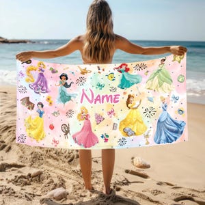 May include: A colourful beach towel featuring Disney princesses, including Cinderella, Belle, Ariel, and Snow White. The towel has a pink, yellow, and blue background with white polka dots and fireworks. The text "Name" is printed in pink on the towel.