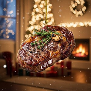 May include: A Christmas ornament shaped like a cooked steak, garnished with rosemary and garlic. The ornament has the name "OLIVIA" printed on it. The background features a Christmas tree and a fireplace.