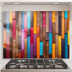 Op de afbeelding: Een kleurrijke houten achterwand met verticale planken in verschillende tinten blauw, oranje, roze, paars en bruin. De planken zijn in een verspringend patroon gerangschikt, waardoor een levendig en gestructureerd oppervlak ontstaat. De achterwand is geïnstalleerd boven een roestvrijstalen gasfornuis.