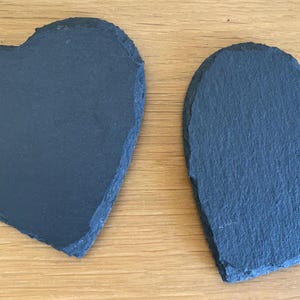 May include: Two heart-shaped slate coasters. The coasters are a dark grey colour with a rough, textured surface. They are placed on a light brown wooden surface.