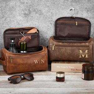 May include: Two brown leather toiletry bags with gold zippers and personalized initials. One bag is open, revealing grooming products. Sunglasses and other grooming products are in front of the bags. The bags are on a wooden surface.