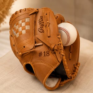 May include: A tan leather baseball glove featuring a woven web pattern, leather lacing, and the name Griffin with the number #18 engraved on the surface. A white baseball with red stitching is tucked inside the pocket of the glove.