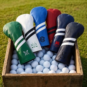 Könnte beinhalten: Eine Sammlung von sechs Golfschlägerhauben aus Leder in Grün, Weiß, Blau, Rot, Marineblau und Schwarz, jeweils mit zwei weißen Streifen und personalisierten Monogrammen oder Symbolen, angeordnet in einer Holzkiste voller weißer Golfbälle.