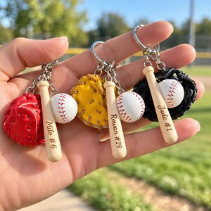 May include: Three baseball-themed keychains featuring miniature gloves in red, yellow, and black, small white baseballs with red stitching, and wooden bats engraved with the names Milo #6, Rowan #19, and Jude #1.