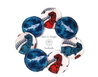 May include: Set of eight round, white cotton makeup remover pads with different colourful printed designs. The designs include a blue and white shark, a red dinosaur with the word "AAAAAH!" and a blue dinosaur with the word "AAAAAH!" The pads are handmade by Josh-n-Ginger.