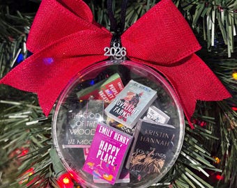 Può includere: Un ornamento trasparente, rotondo e piatto riempito con repliche di libri in miniatura, inclusi titoli come Happy Place, The Four Winds e Lovelight Farms. Un fiocco in tessuto rosso è posto in cima con un ciondolo argentato che mostra l'anno 2026.