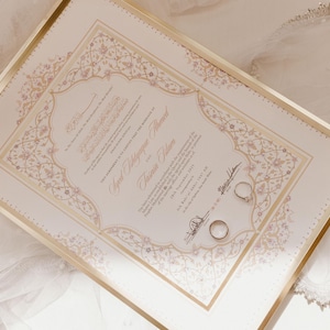Könnte beinhalten: Eine gerahmte Hochzeitsurkunde mit goldenen und floralen Mustern. Zwei Eheringe liegen auf der Urkunde. Ein dekorativer Kopfschmuck und Schleier befinden sich im Hintergrund.