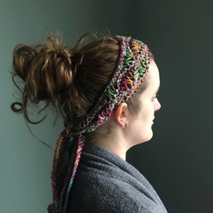 May include: A woman with brown hair wearing a colourful crocheted headband. The headband is tied in a knot at the back of her head and the ends are hanging down her back.