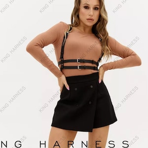 May include: A woman wearing a brown long-sleeved shirt with a black leather harness and a black mini skirt. The text "KING HARNESS MATURE" is repeated in the background.