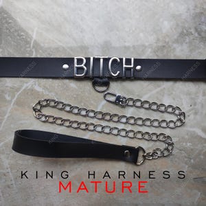 Puede incluir: Gargantilla de cuero negro con la palabra "BITCH" en letras plateadas. Incluye una correa de cadena plateada y un asa. El texto "KING HARNESS" y "MATURE" está impreso en la parte inferior.