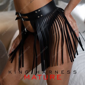 Puede incluir: Falda de flecos de cuero negro con cinturón de hebilla y tachuelas plateadas. La falda está hecha de cuero sintético y tiene una cintura alta.