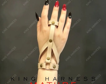 May include: A beige leather finger and wrist harness with a ring and multiple straps. The harness has a decorative metal stud on each strap.