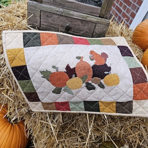 Op de afbeelding: Een gewatteerde placemat met herfstthema met pompoenen, bladeren en een eekhoorn. De rand heeft een patchworkpatroon in herfstkleuren. De placemat ligt op een bed van hooi, met pompoenen op de achtergrond, wat een seizoensgebonden display creëert.
