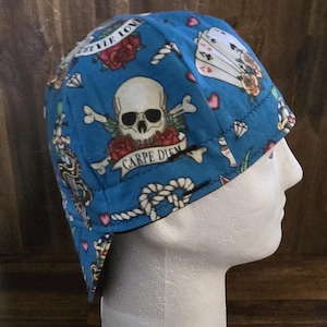 May include: Blue welding cap with a repeating tattoo-style design. The cap features a skull with crossed bones, roses, diamonds, playing cards, and nautical rope patterns. The words "Forever Love" and "Carpe Diem" are also visible.