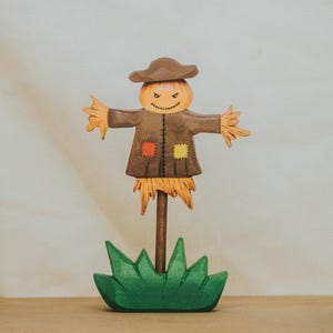 May include: A wooden scarecrow figurine featuring a brown hat, a brown coat with orange and yellow patches, and a stitched face. The figure stands on a green base with a central brown pole, displaying straw-like details at the sleeves and hem.