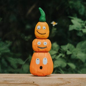 Könnte beinhalten: Ein vertikaler Stapel aus drei orangefarbenen kürbisförmigen Figuren. Der oberste Kürbis hat einen grünen Stiel und einen genähten Mund. Der mittlere Kürbis zeigt ein breites Lächeln. Der untere Kürbis hat einen überraschten Gesichtsausdruck.