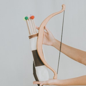 May include: A wooden bow and arrow set. The bow is light brown, and the quiver is white with a brown stripe and the text "Kataring". The arrows have colourful pom-poms on the tips. The set is held in front of a white background.