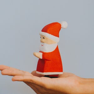 以下が含まれることがあります： 人の手に持たれた、白いひげと帽子をつけた赤い小さなサンタクロースの置物です