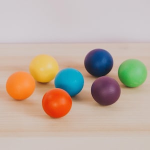 May include: Seven colorful wooden balls in various shades of red, orange, yellow, green, blue, and purple. The balls are arranged on a light wood surface.