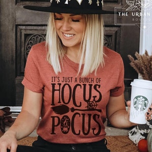 Könnte beinhalten: Eine Frau trägt ein rostfarbenes T-Shirt mit dem Text "It's just a bunch of Hocus Pocus" in Schwarz. Das Shirt zeigt ein schwarzes Besen- und Spinnennetz-Motiv. Sie hält eine weiße Kaffeetasse mit einem schwarzen Logo.