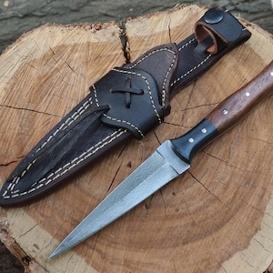 May include: A handmade dagger with a patterned blade and a wooden handle, resting on a wooden surface. The dagger is accompanied by a dark brown leather sheath with white stitching and a belt loop.