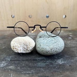 May include: Black round-frame spectacles with a bridge and nose pads, resting on a grey stone. The spectacles have a minimalist design. A white coral-like object is to the left of the stone. The background is a wooden surface.