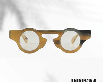 Könnte beinhalten: Rundbrillen mit einem einzigartigen Hornrand-Design. Die Fassungen sind eine Mischung aus Braun- und Beigetönen mit klaren Gläsern. Die Brille hat eine markante Form, mit dem Markennamen "PRISM" unten.