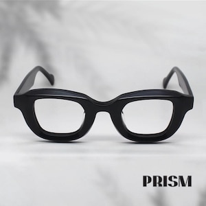 May include: Black eyeglasses with a thick, rectangular frame. The glasses have a bold, modern design with clear lenses. The brand name "PRISM" is printed in a bold, sans-serif font at the bottom of the image.
