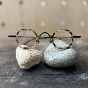 May include: Round eyeglasses with a black metal frame and tortoise shell patterned rims. The glasses are displayed on a gray stone and a white rock. The lenses are clear. The overall design is vintage and stylish.