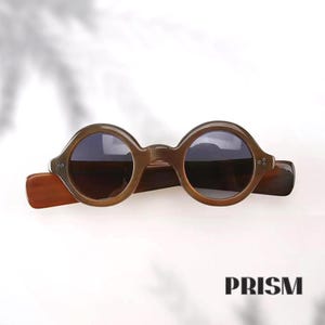 May include: Round, brown sunglasses with dark lenses. The sunglasses have a thick frame and arms. The brand name "PRISM" is printed in black at the bottom right corner of the image.