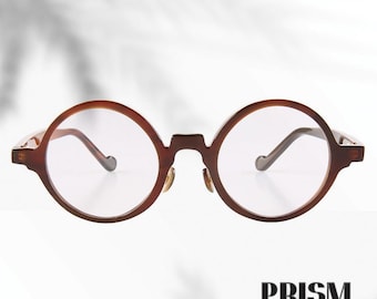 Könnte beinhalten: Runde, braune Brille mit klaren Gläsern. Die Fassungen haben eine klassische, kreisförmige Form und einen Schlüssellochsteg. Die Bügel sind dünn und biegen sich sanft. Das Wort "PRISM" ist unten in fetter Schrift gedruckt.