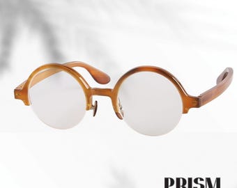 Könnte beinhalten: Runde, braune Brille mit klassischem Design. Die Fassungen sind in einem warmen Bernsteinton gehalten, die Gläser sind klar. Die Brille hat einen Schlüssellochsteg und ist für den täglichen Gebrauch geeignet. Das Wort "PRISM" ist unten in Schwarz gedruckt.