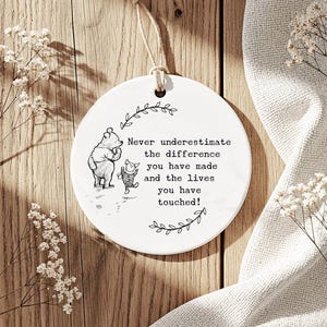 May include: A circular white ceramic decoration featuring Winnie the Pooh and Piglet illustrations and a quote. The text reads, "Never underestimate the difference you have made and the lives you have touched!" A jute string is attached for hanging.