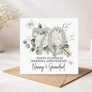 May include: A white card with a silver glitter "70" and the text "Happy Platinum Wedding Anniversary Nanny & Grandad". The card features white roses, greenery, and butterflies. A small tag reads "1956-2026". The card is on a brown envelope.